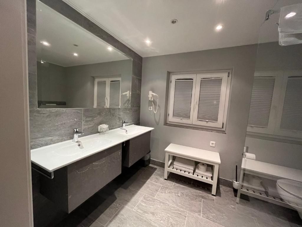 A modern bathroom with a double sink vanity, a bench, and a toilet.