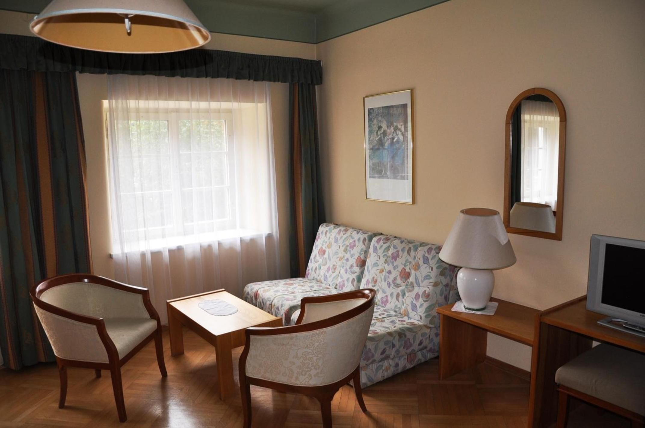 A room with a sofa, two chairs, a table, a lamp, a TV, and a window with curtains.