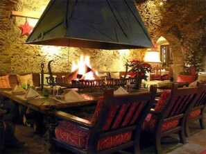 Un coin repas rustique avec un mur en pierre, une grande cheminée, des tables en bois et des chaises à motifs rouges.