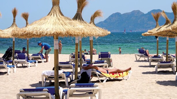 A beach with sun loungers and thatched umbrellas, a clear blue sea, and distant mountains. - Hoposa Cultural Boutique (Pollenca)