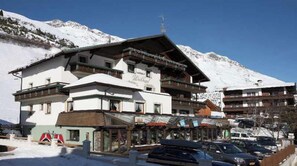 A multi-story hotel with balconies, a ski slope in the background, and a parking area.