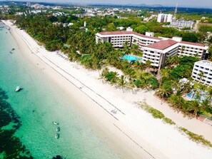 An aerial view of a beach resort with a white sandy beach, clear turquoise waters, and a large building with multiple balconies.