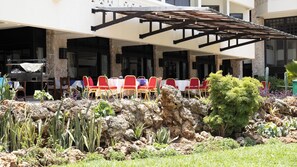 Un espace extérieur aménagé pour les repas, avec des chaises rouges et jaunes, une terrasse couverte et un jardin de rocaille agrémenté de différentes plantes.