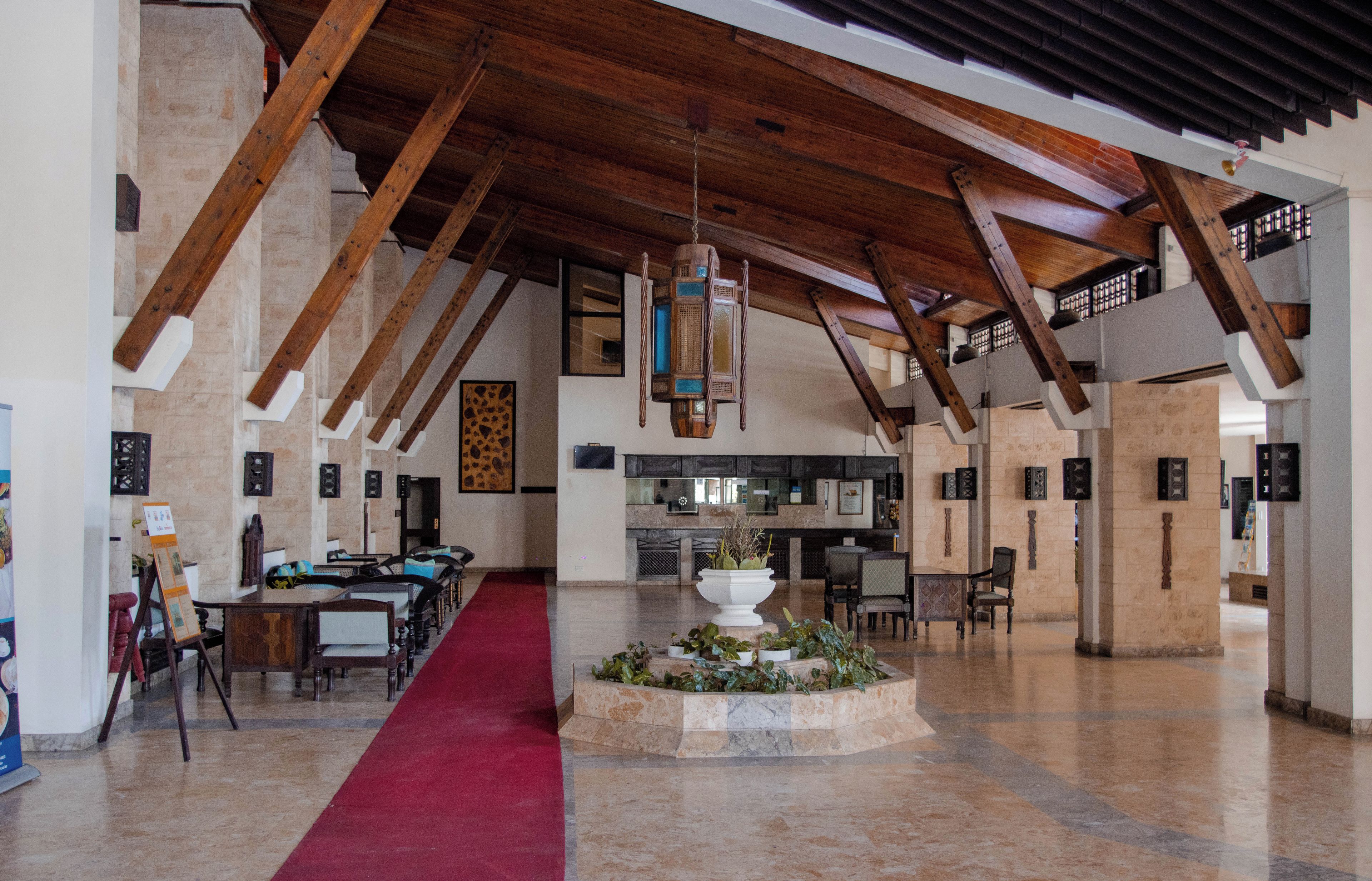 Un hall espacioso con altos vigas de madera, una alfombra roja central y un área de descanso con mesas y sillas.