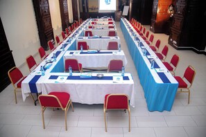Une salle de conférence avec de longues tables, des chaises rouges et un écran de projection à l’avant.