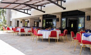 An outdoor dining area with tables and chairs, stone flooring, and a covered section with a pergola.
