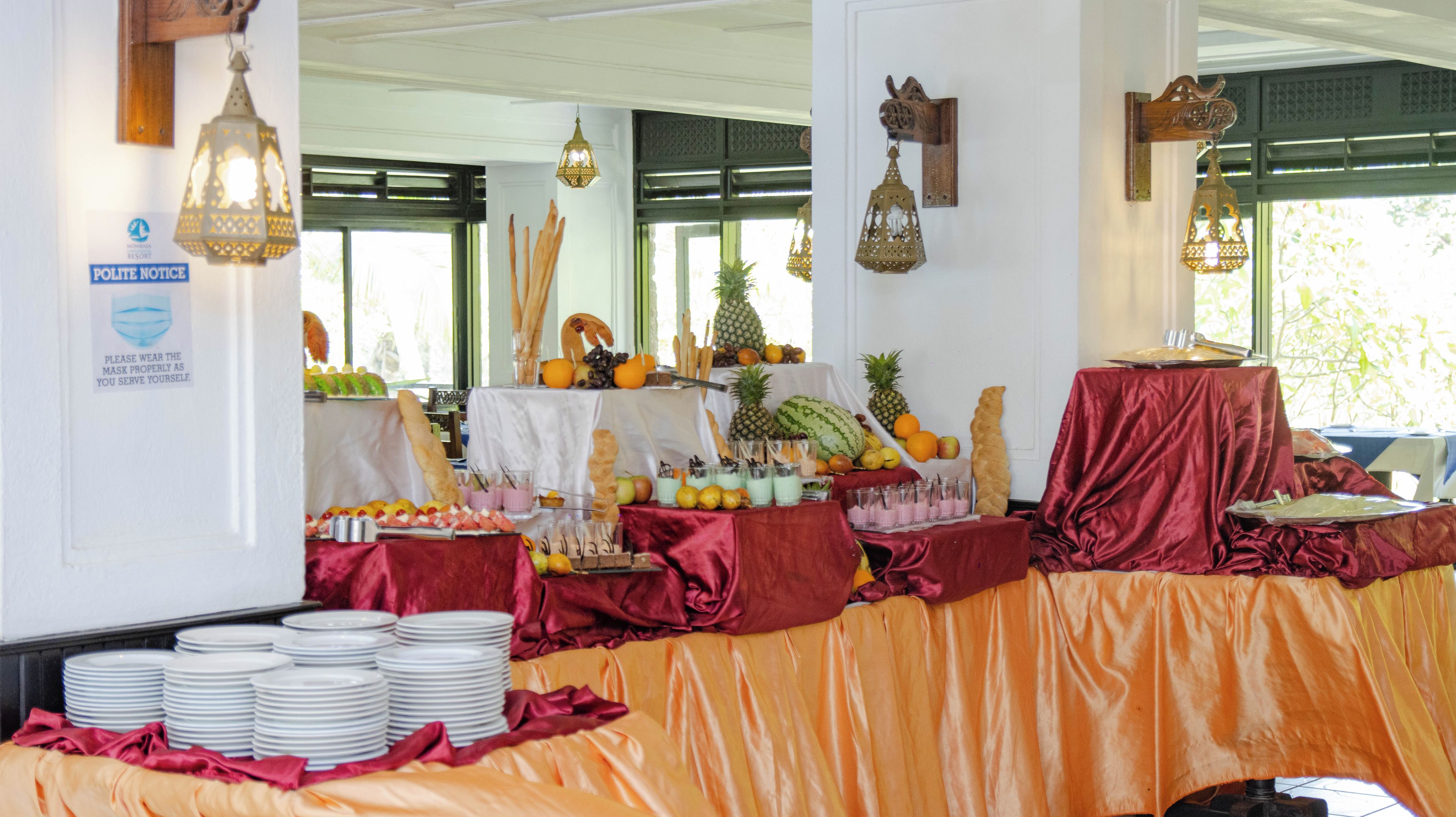 A buffet setup with various fruits, pastries, and beverages on tables covered with red and orange cloths.