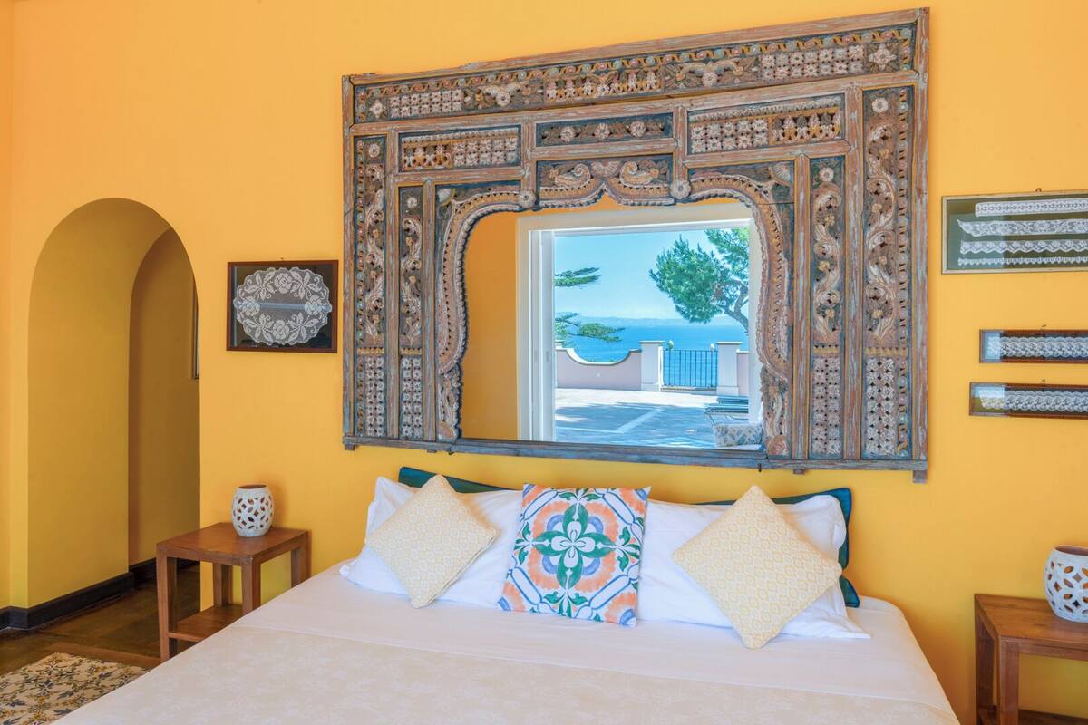 A room with a large bed, a decorative mirror, and a view of the sea.
