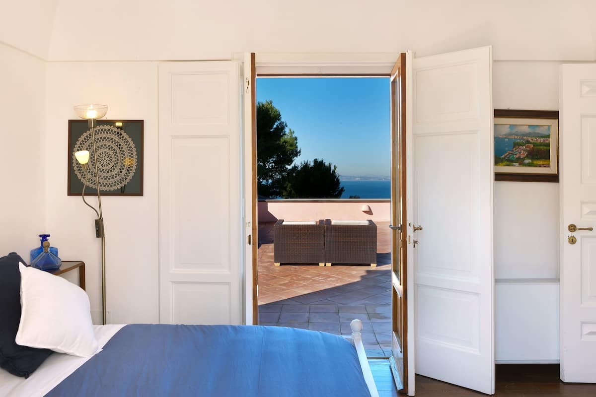 A bedroom with a bed, a nightstand, a lamp, and a view of an outdoor patio with furniture.