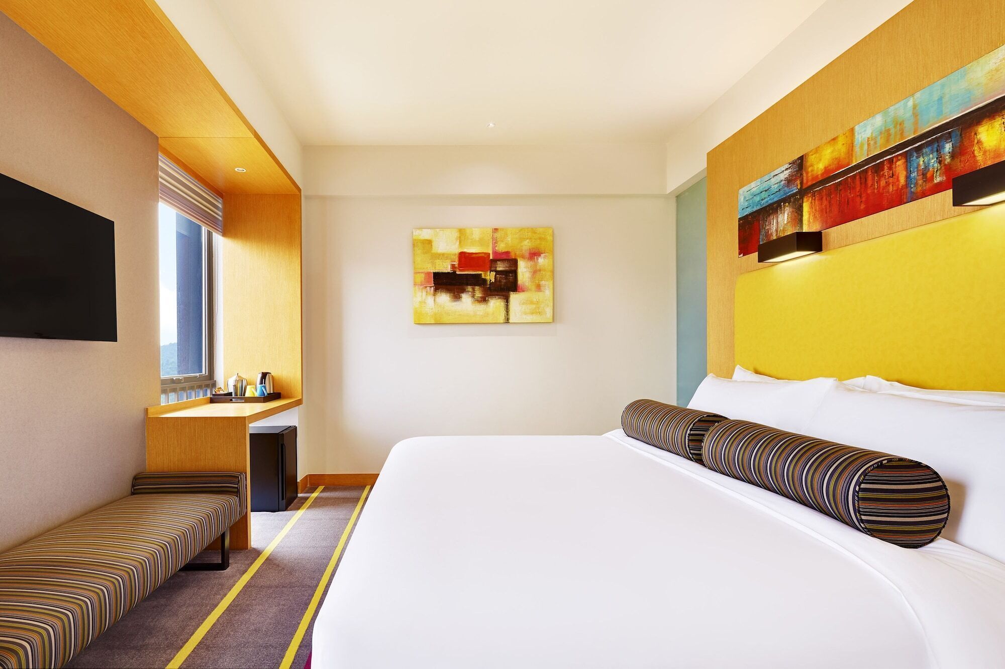 A hotel room with a bed, a bench, a desk, a TV, and a painting on the wall.
