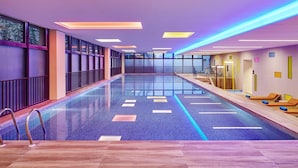 A large indoor swimming pool with a tiled floor, surrounded by walls with large windows, and a wooden deck area with lounge chairs. - Aloft Dongguan Dynamic Town (Shenzhen)