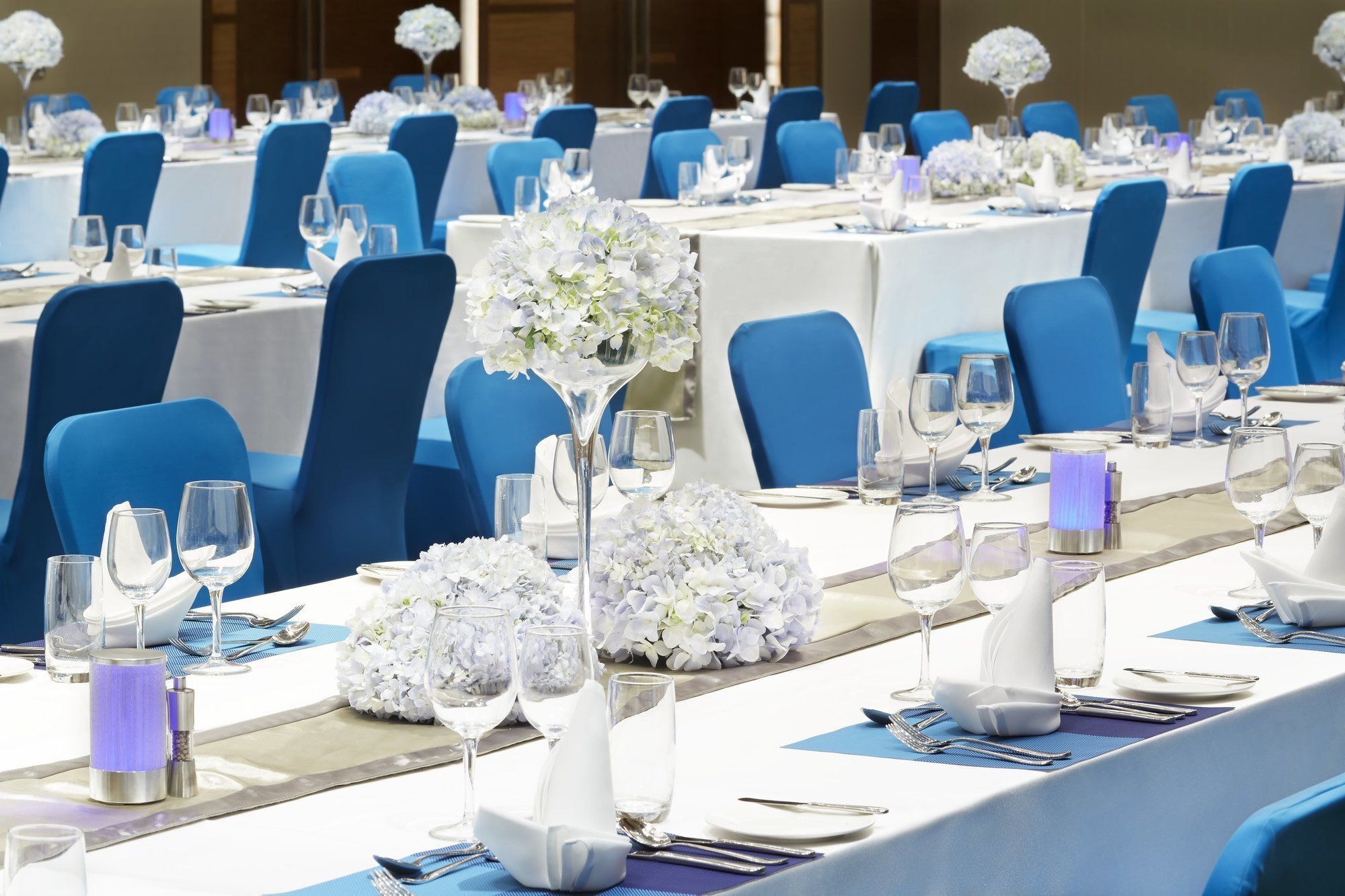 A banquet hall with tables set for a formal event, featuring blue chairs, white tablecloths, and floral centerpieces.