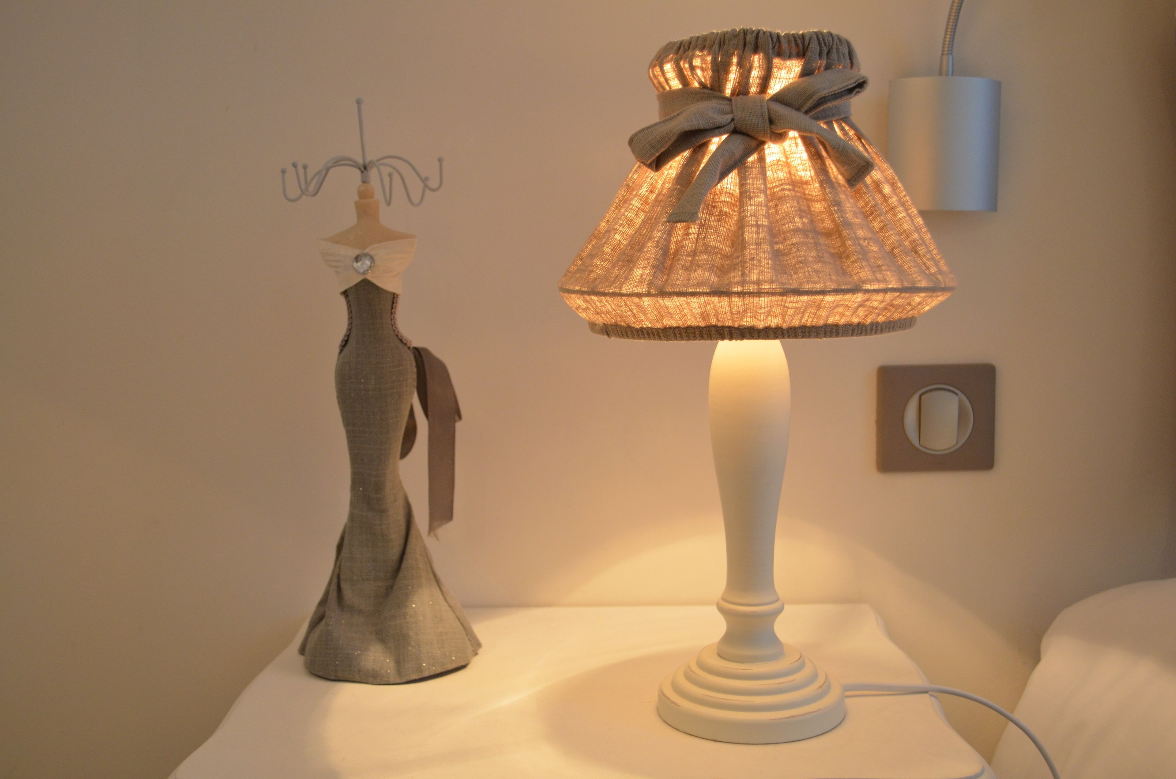 A lamp with a textured shade and a bow on a bedside table next to a wall-mounted light fixture.