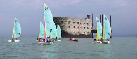 A group of sailboats with numbered sails, a historic stone building, and calm waters.
