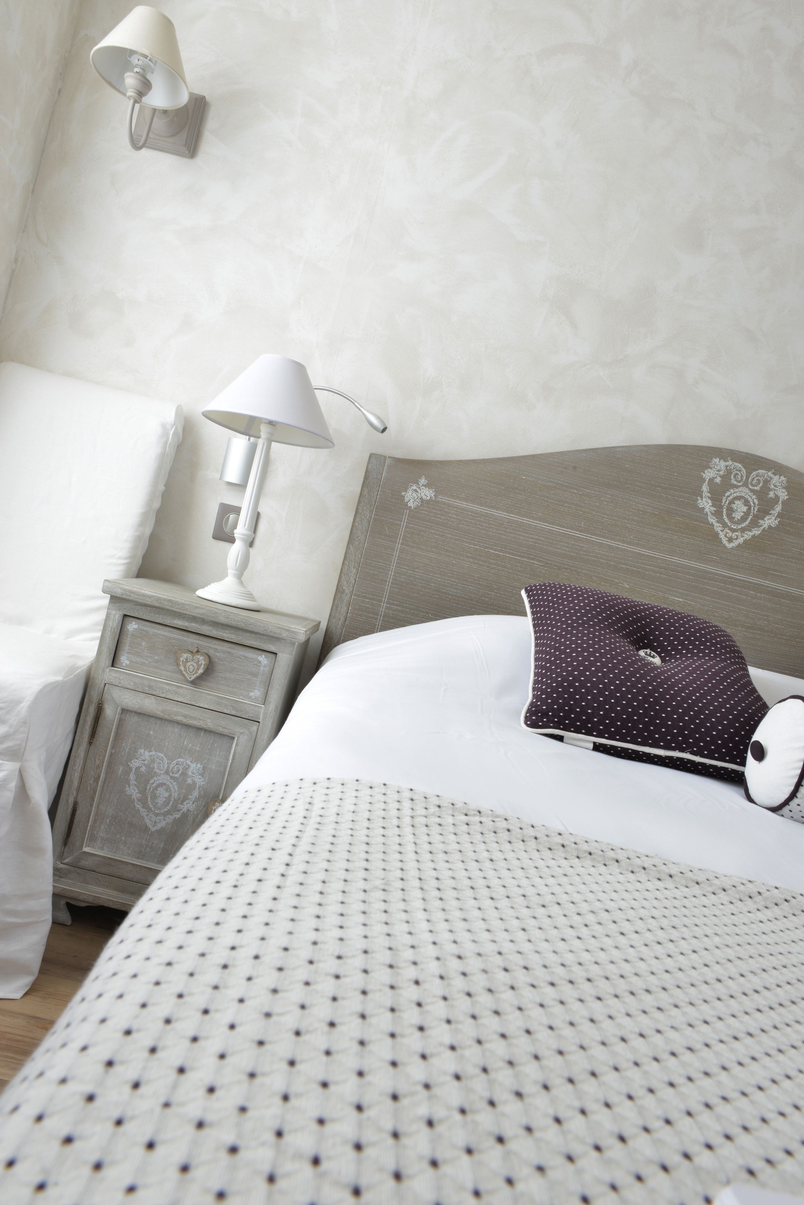 A neatly made bed with a patterned bedspread, a heart-shaped drawer on the nightstand, a white lamp, and a decorative pillow.