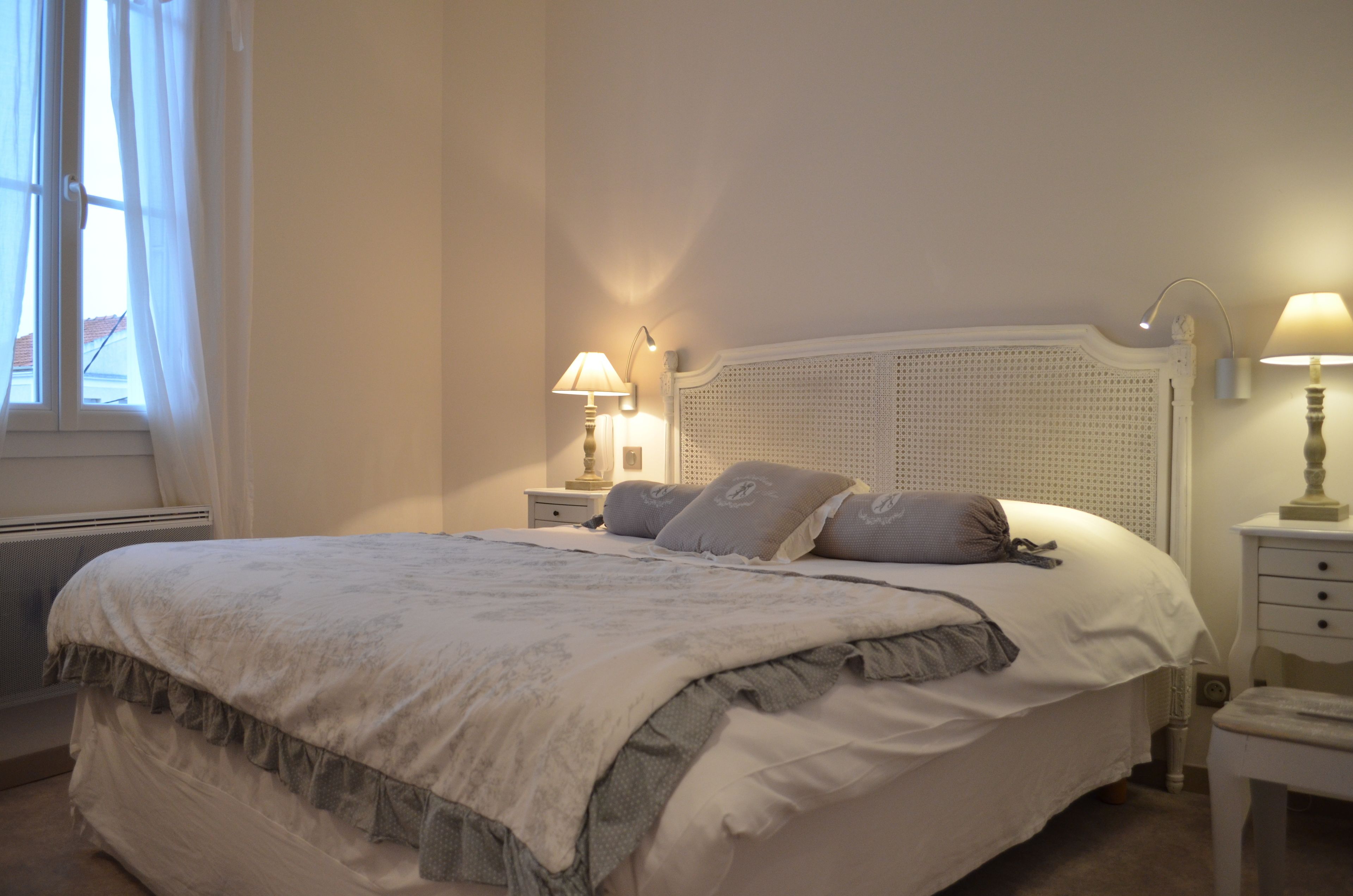 Een netjes opgemaakt bed met een wit hoofdeinde, twee nachtkastjes met lampen en een raam met gordijnen.