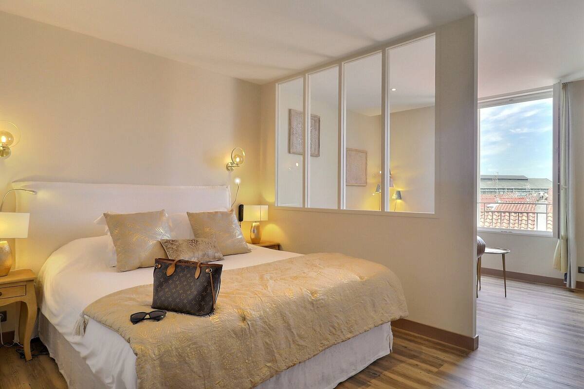 Une chambre bien éclairée, dotée d’un grand lit, de lampes de chevet et offrant une vue sur la ville, ainsi qu’un balcon.