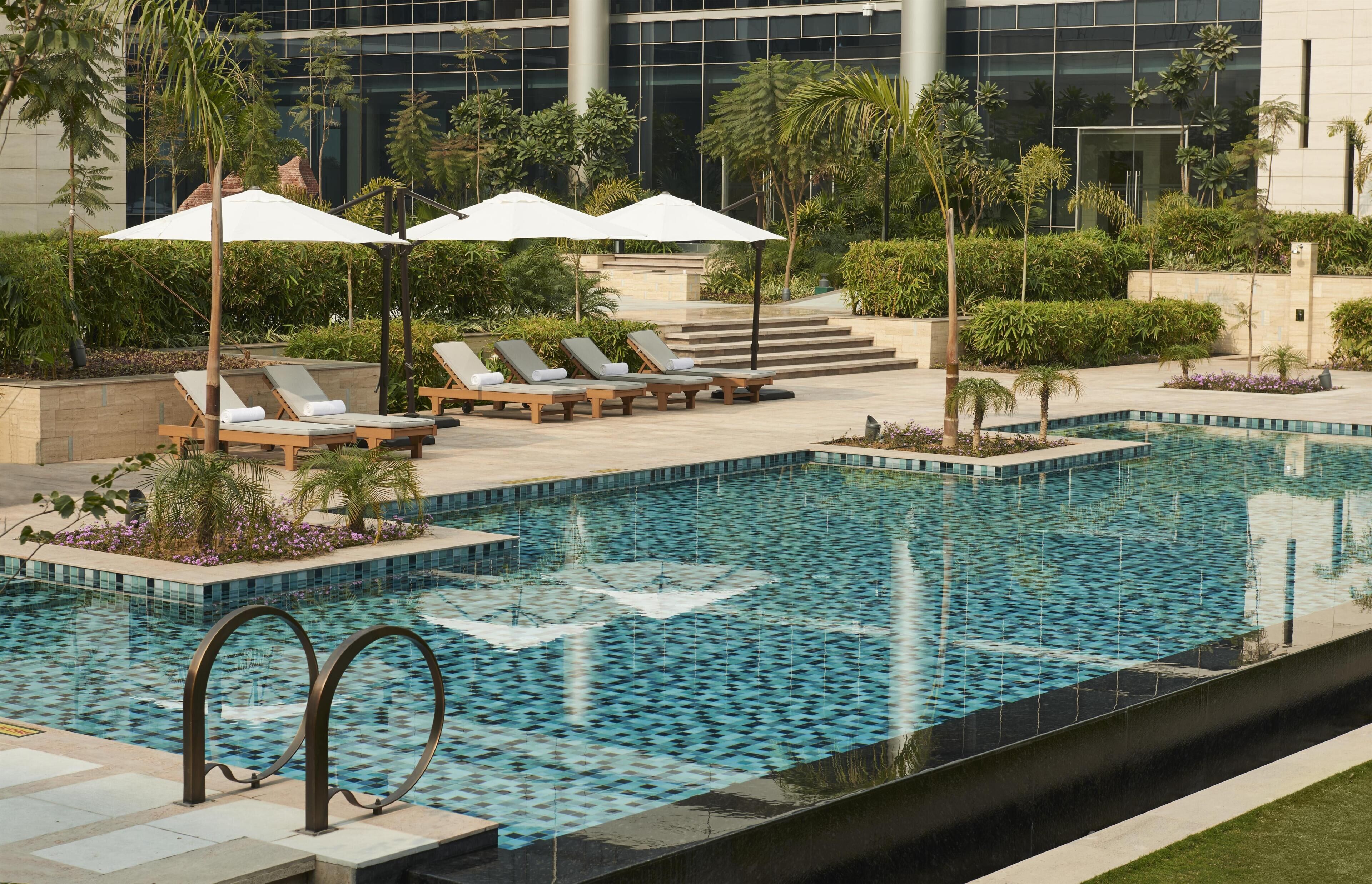 A pool area with lounge chairs and umbrellas, surrounded by greenery and a modern building in the background.