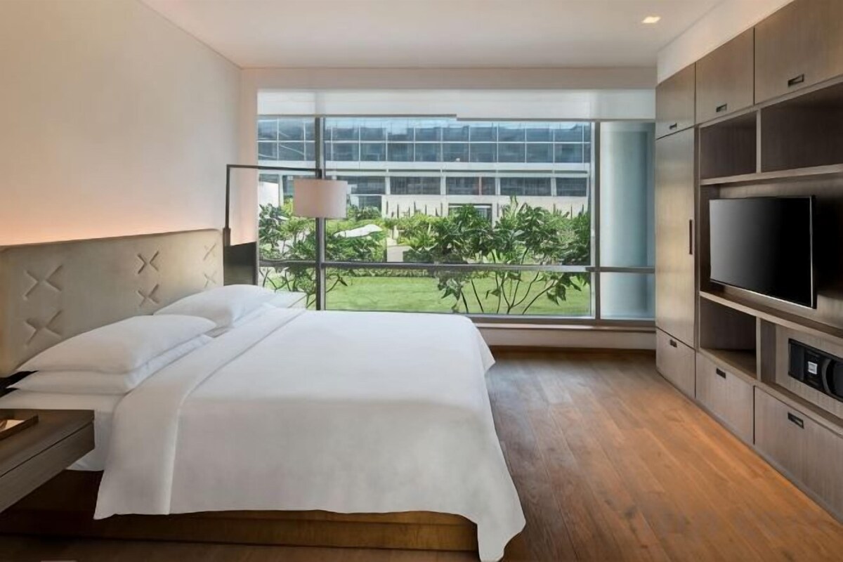 A modern hotel room with a large bed, a TV, and a view of greenery outside.