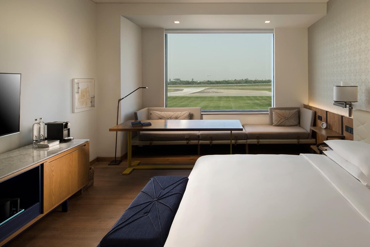 A hotel room with a large bed, a desk with a laptop, a sofa, a window with a view of the airport, and a TV on the left.