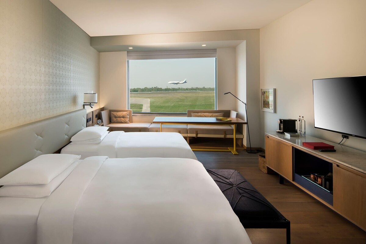 A hotel room with two beds, a desk, a TV, and a window with an airplane view.