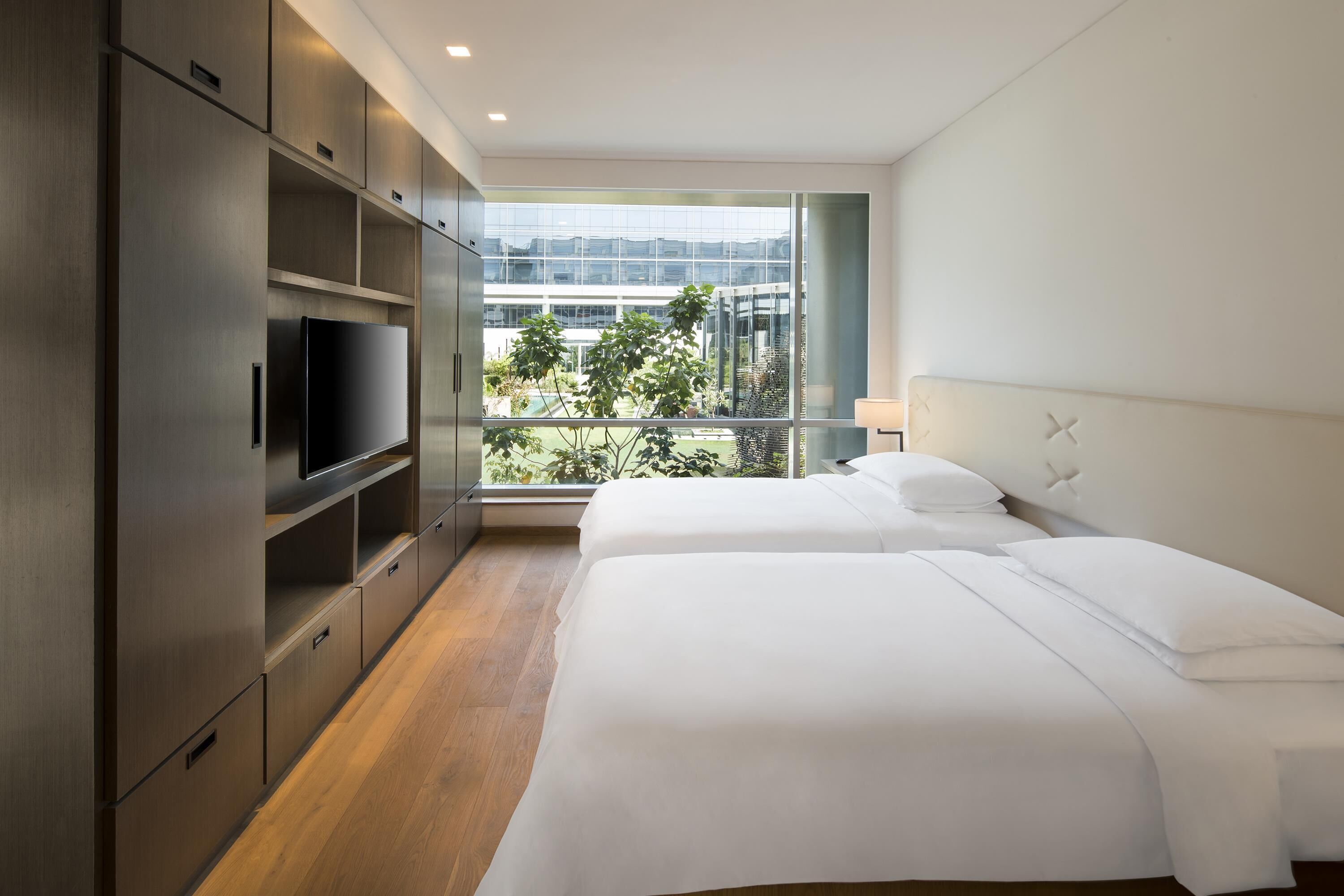 Habitación de hotel con cama, televisor, ventana grande y vistas a un edificio y vegetación.