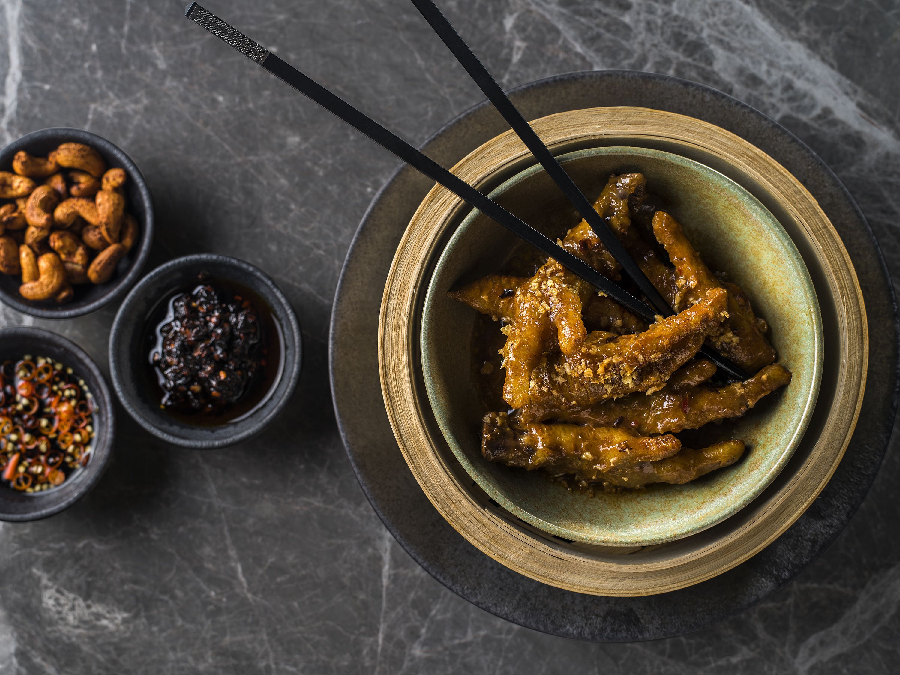 A bowl of glazed chicken wings with black chopsticks, accompanied by small bowls of nuts and sauces on a dark surface.
