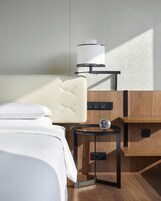 Una habitación de hotel ordenada con una cama, una mesita de noche con lámpara y una mesita auxiliar de madera.