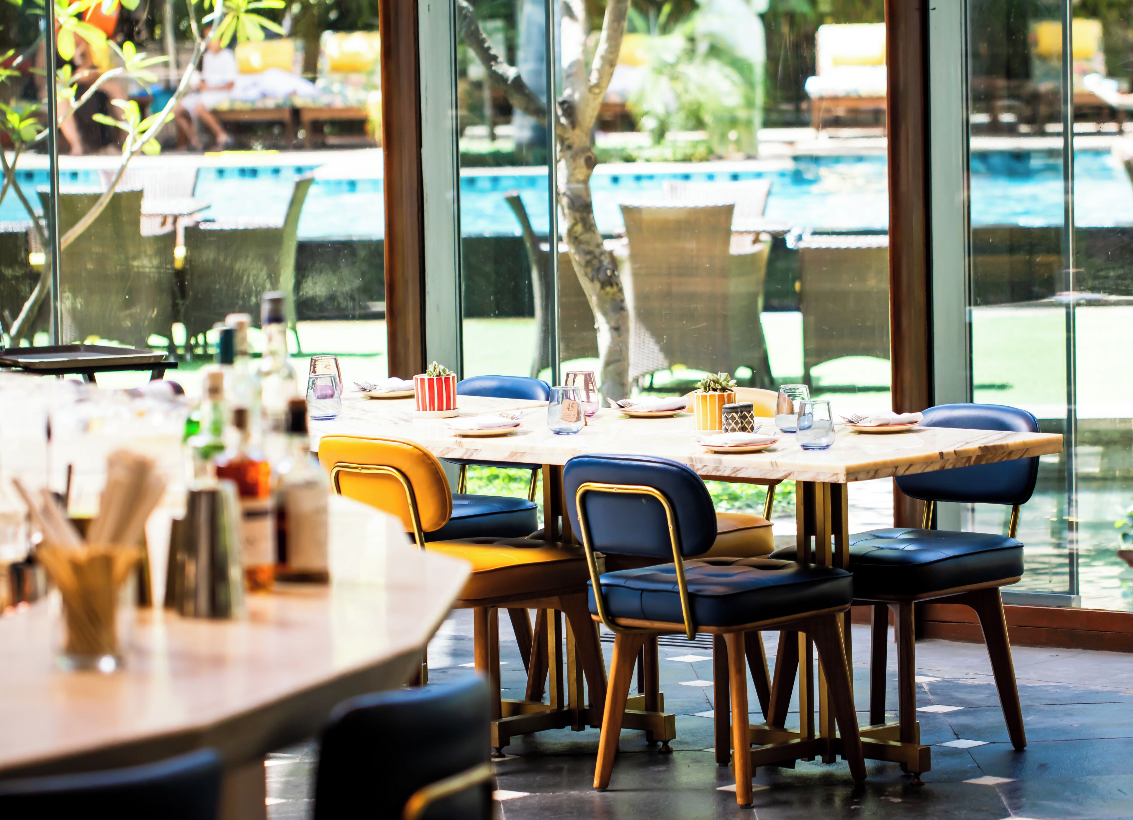 A dining area with a pool view, tables set with plates, glasses, and cutlery, and chairs with cushioned seats.