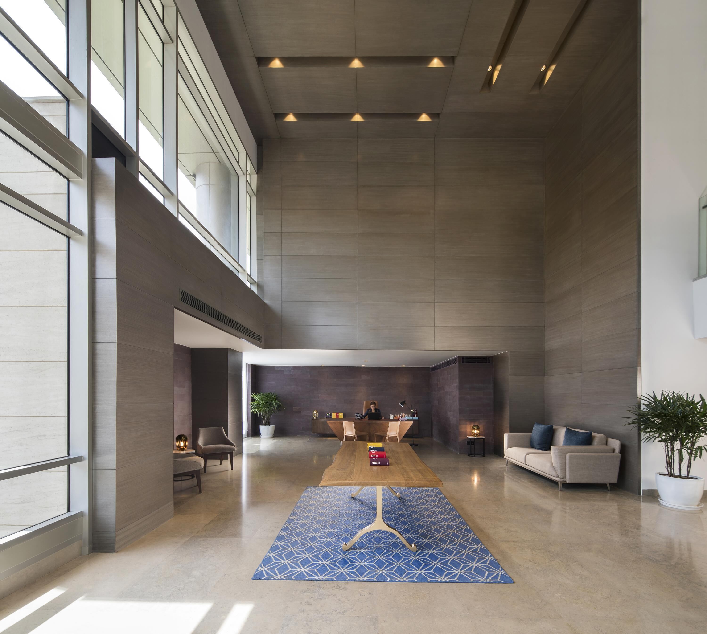 A spacious lobby with a high ceiling, a central wooden table, seating areas, and large windows allowing natural light.
