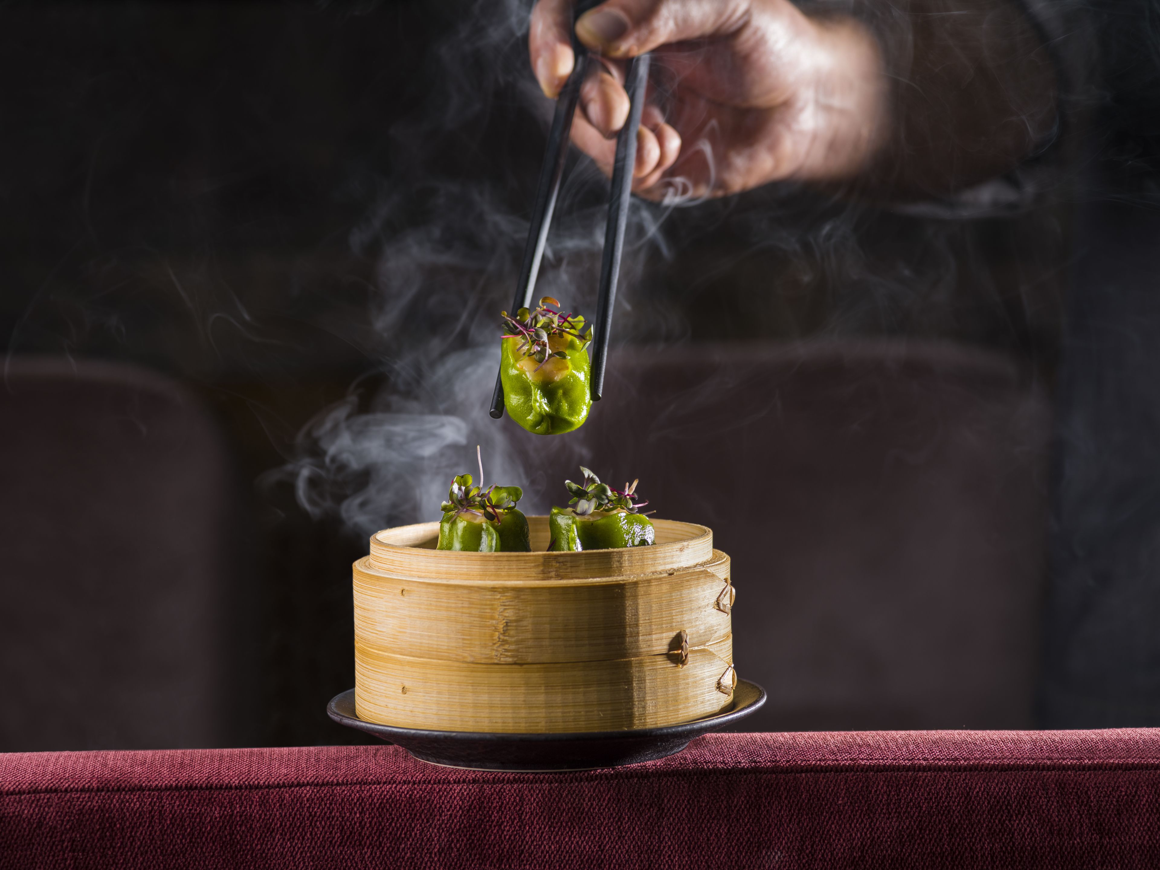 A bamboo steamer with food items, steam rising, and a hand holding tongs.