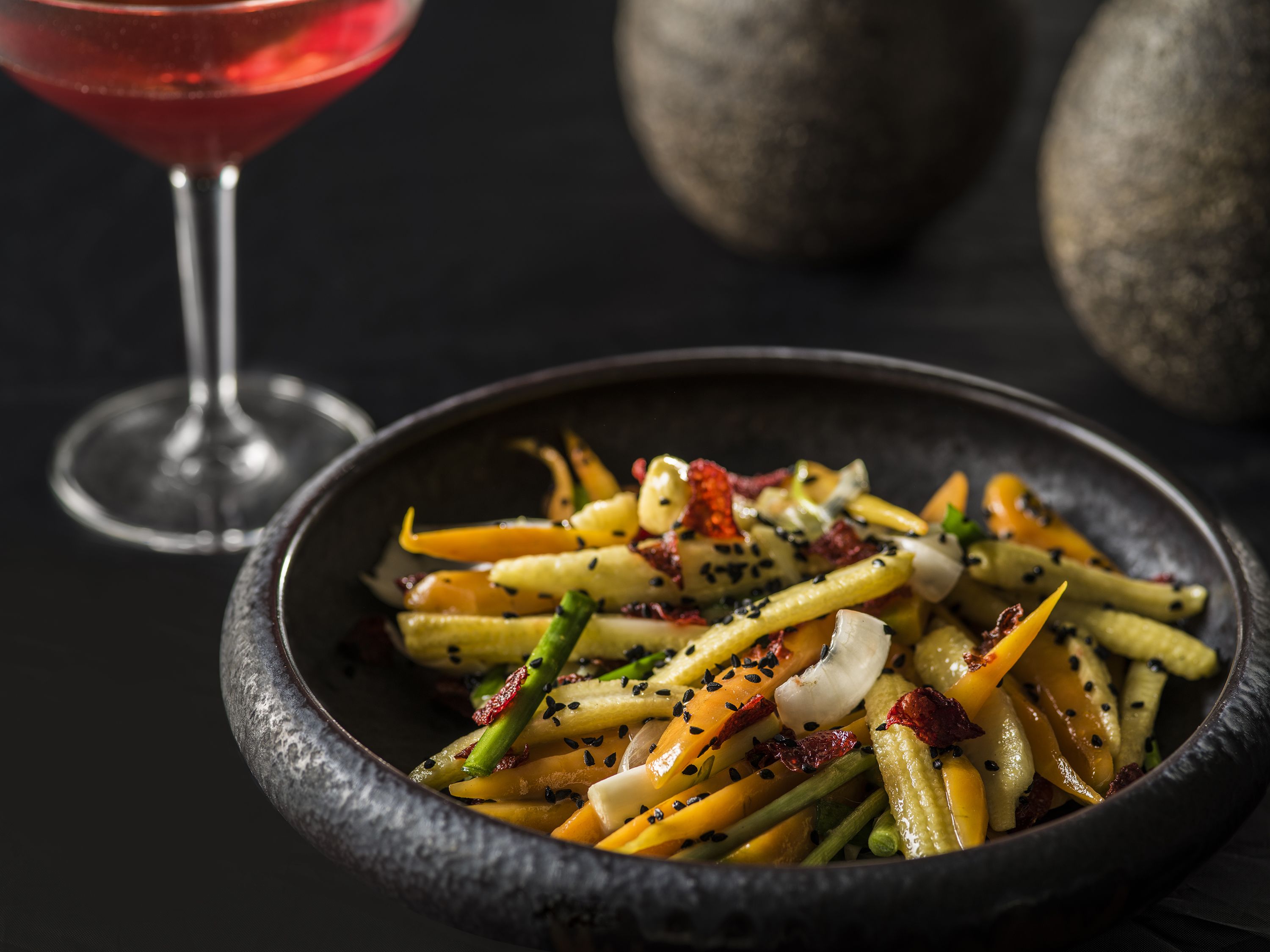 A bowl of colorful vegetable salad with sesame seeds, accompanied by a glass of red wine and a dark bowl.