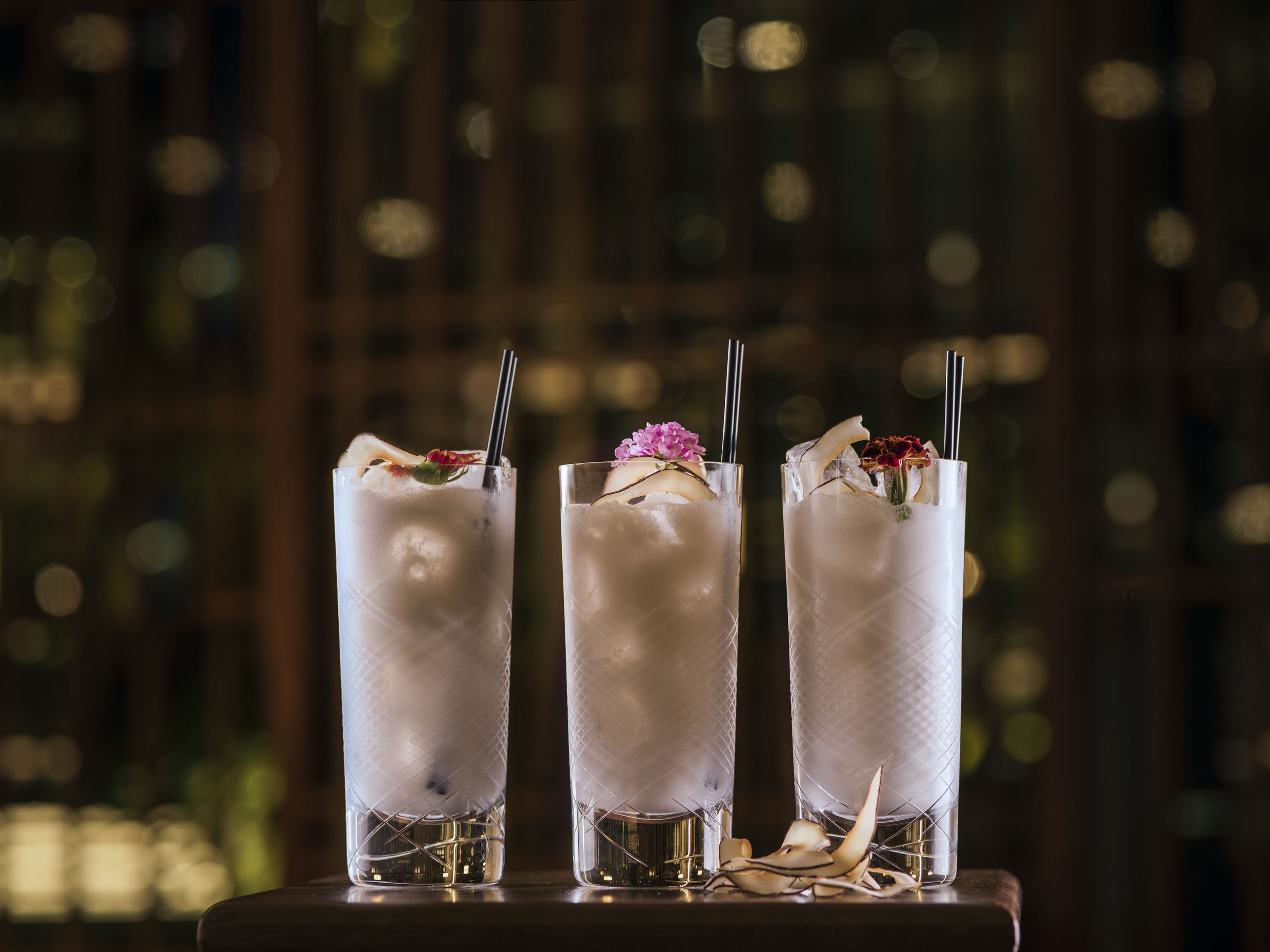 Three tall glasses of a clear beverage with ice, garnished with flowers and a straw, placed on a wooden surface.