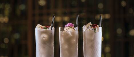 Three tall glasses of a clear beverage with ice, garnished with flowers and a straw, placed on a wooden surface.
