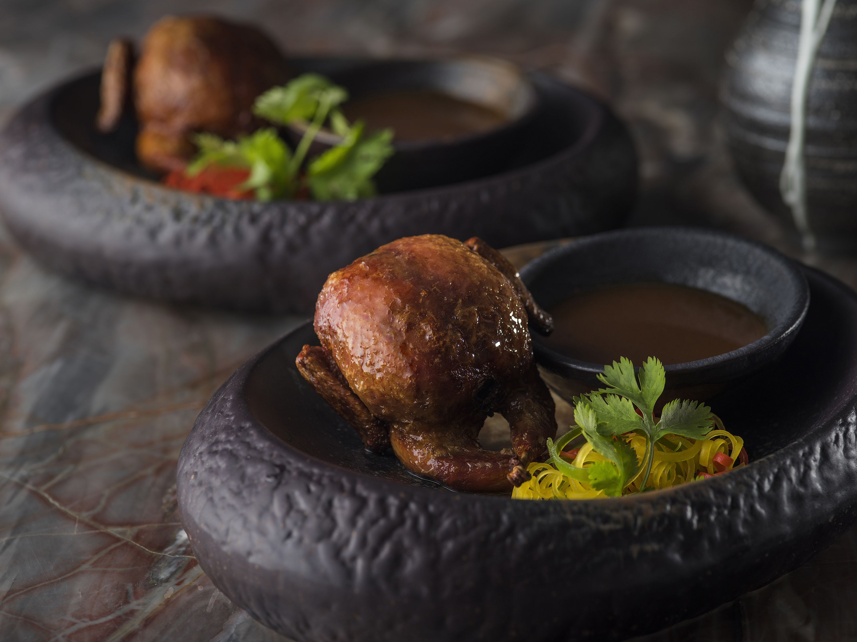 A roasted duck in a rustic, dark bowl with a side of sauce and garnish.