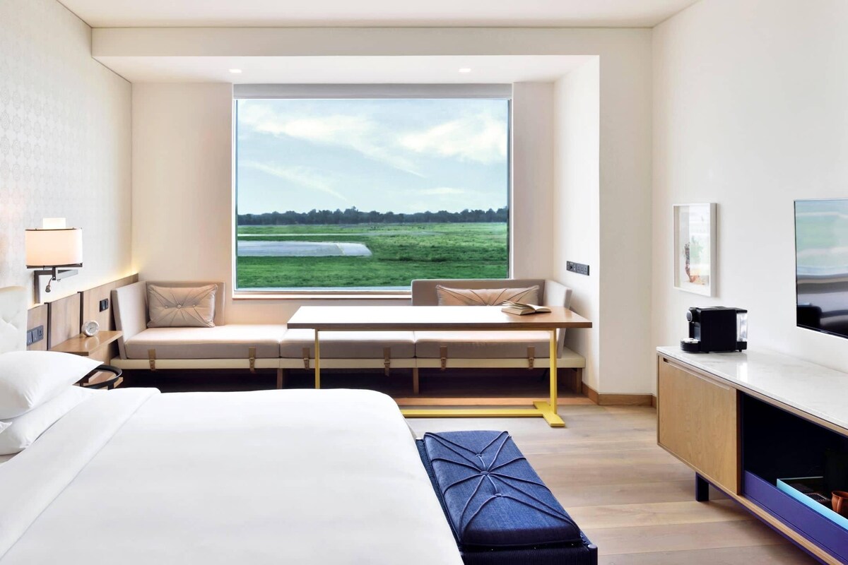 A hotel room with a large bed, a seating area with a view of a green landscape, a desk with a lamp, and a television.