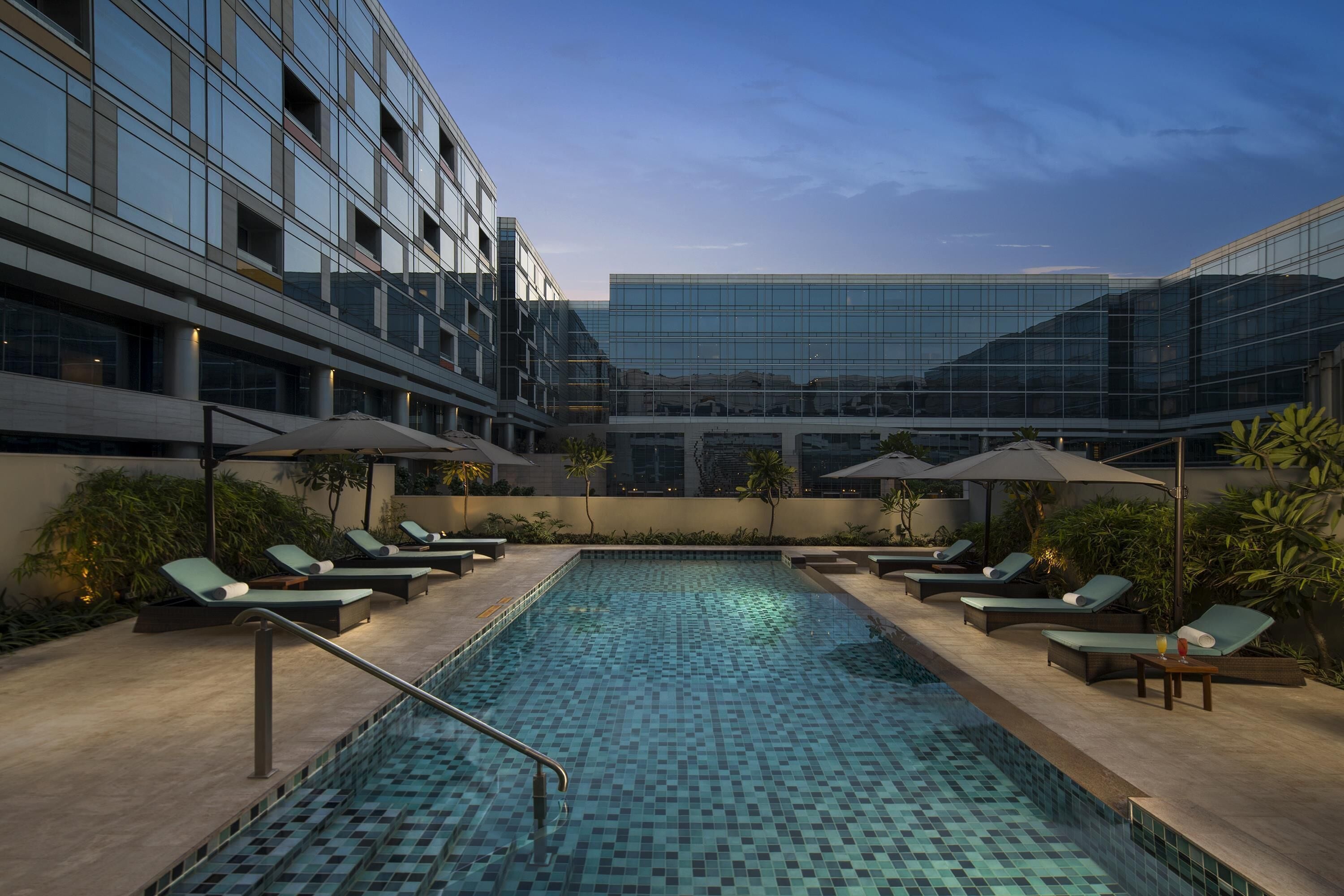 A modern indoor pool with lounge chairs and umbrellas, surrounded by glass buildings.
