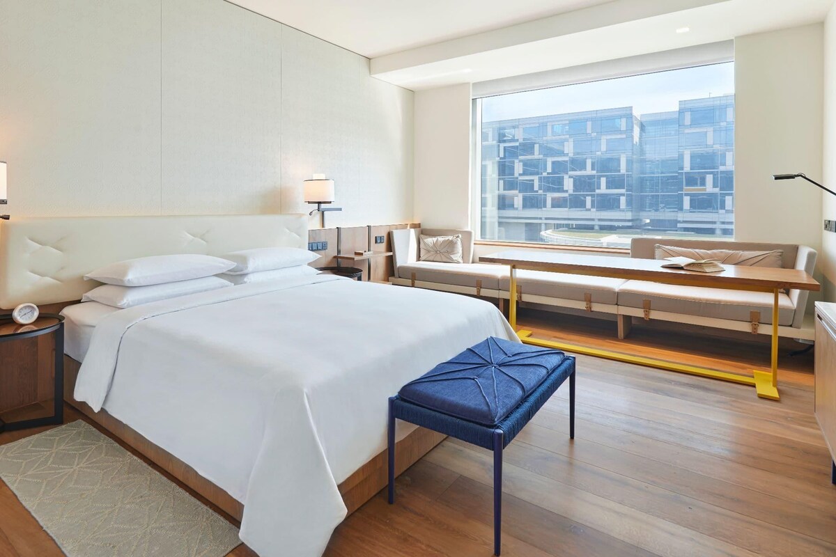 A modern hotel room with a large bed, bedside tables, a reading area with a bench, and a large window offering a city view.