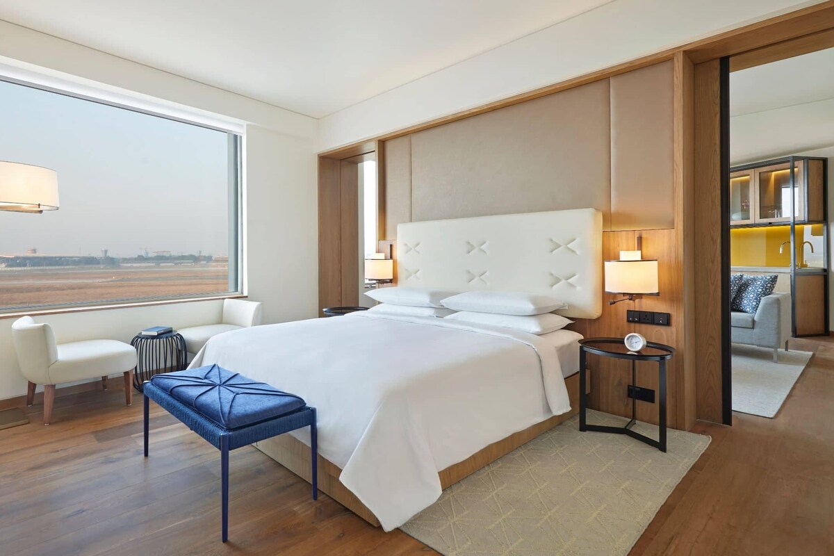 A modern hotel room with a large bed, a sitting area with a chair and a small table, a bedside lamp, and a view of the airport.