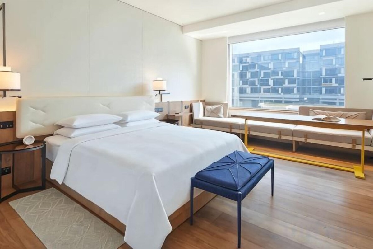 A hotel room with a large bed, bedside tables, a bench, a window with a view of buildings, and a sofa.