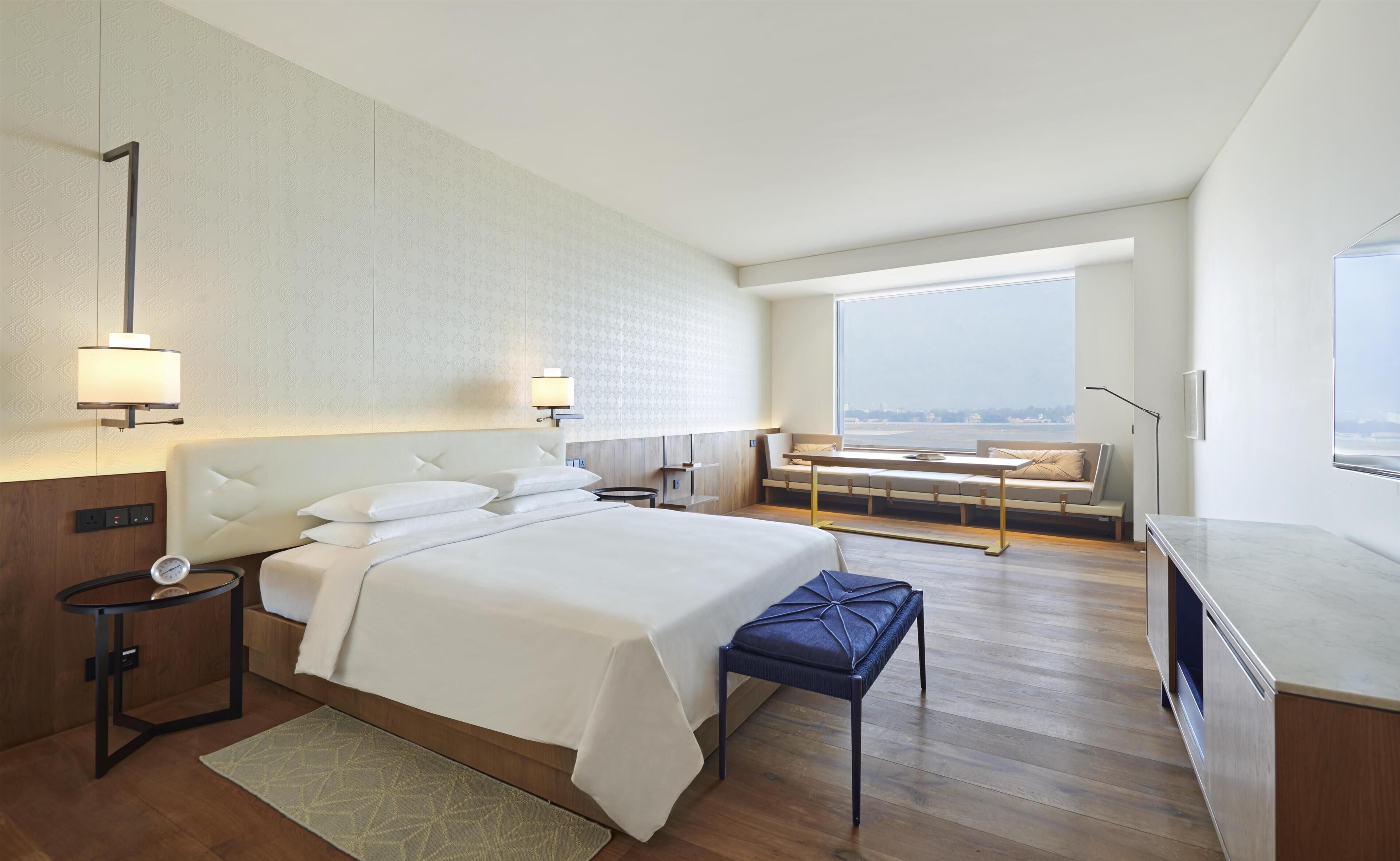 A modern hotel room with a large bed, bedside tables, a seating area with a view, and a large window.