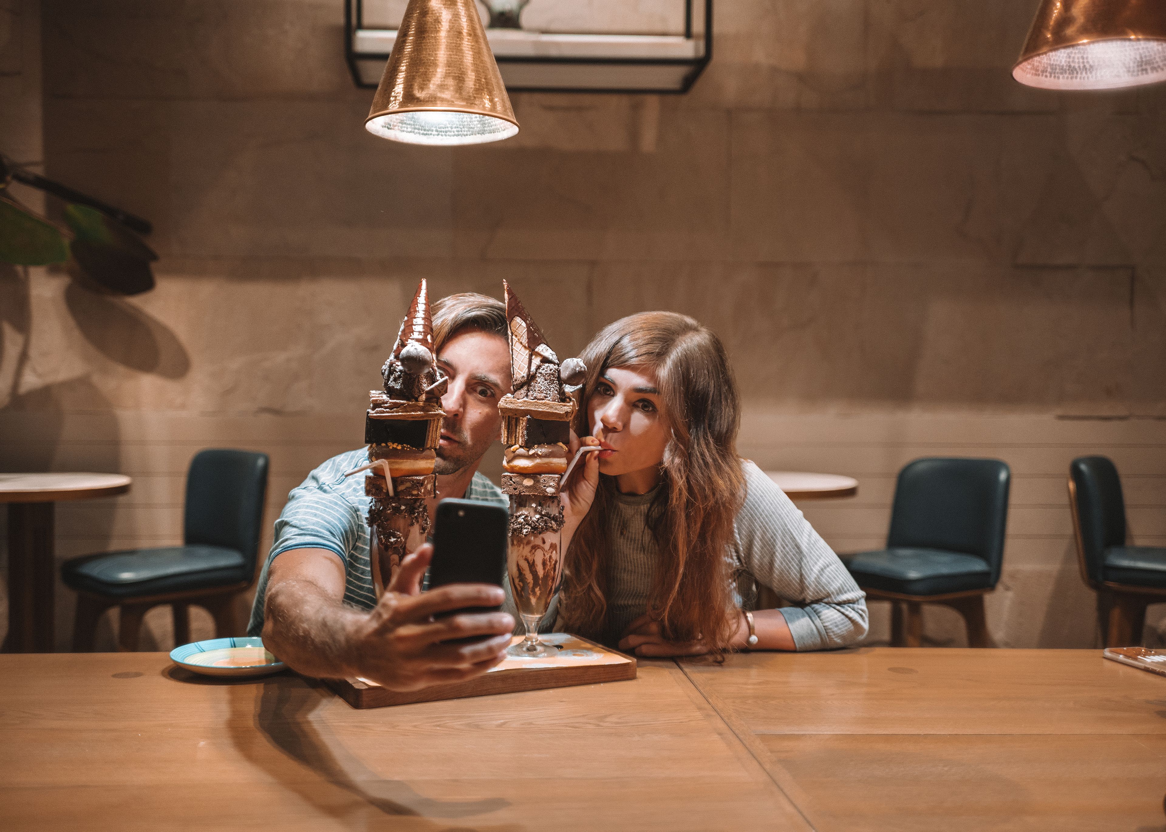 A couple taking a selfie at a wooden table in a cafe with a modern interior, including a hanging light fixture and cushioned chairs.