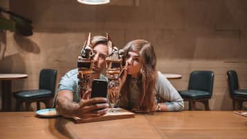 A couple taking a selfie at a wooden table in a cafe with a modern interior, including a hanging light fixture and cushioned chairs.