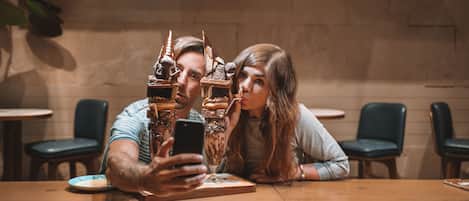 Ein Paar macht ein Selfie an einem Holztisch in einem Café mit modernem Interieur, inklusive einer Hängelampe und gepolsterten Stühlen.