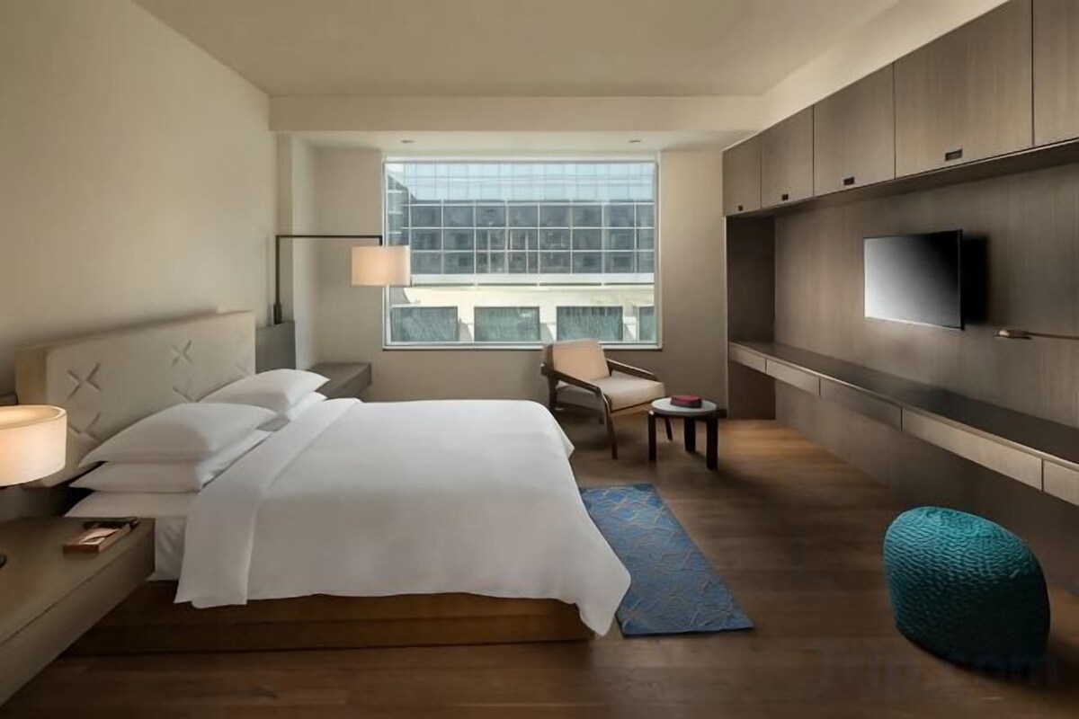 A hotel room with a bed, bedside table, lamp, chair, TV, and a window with a view.