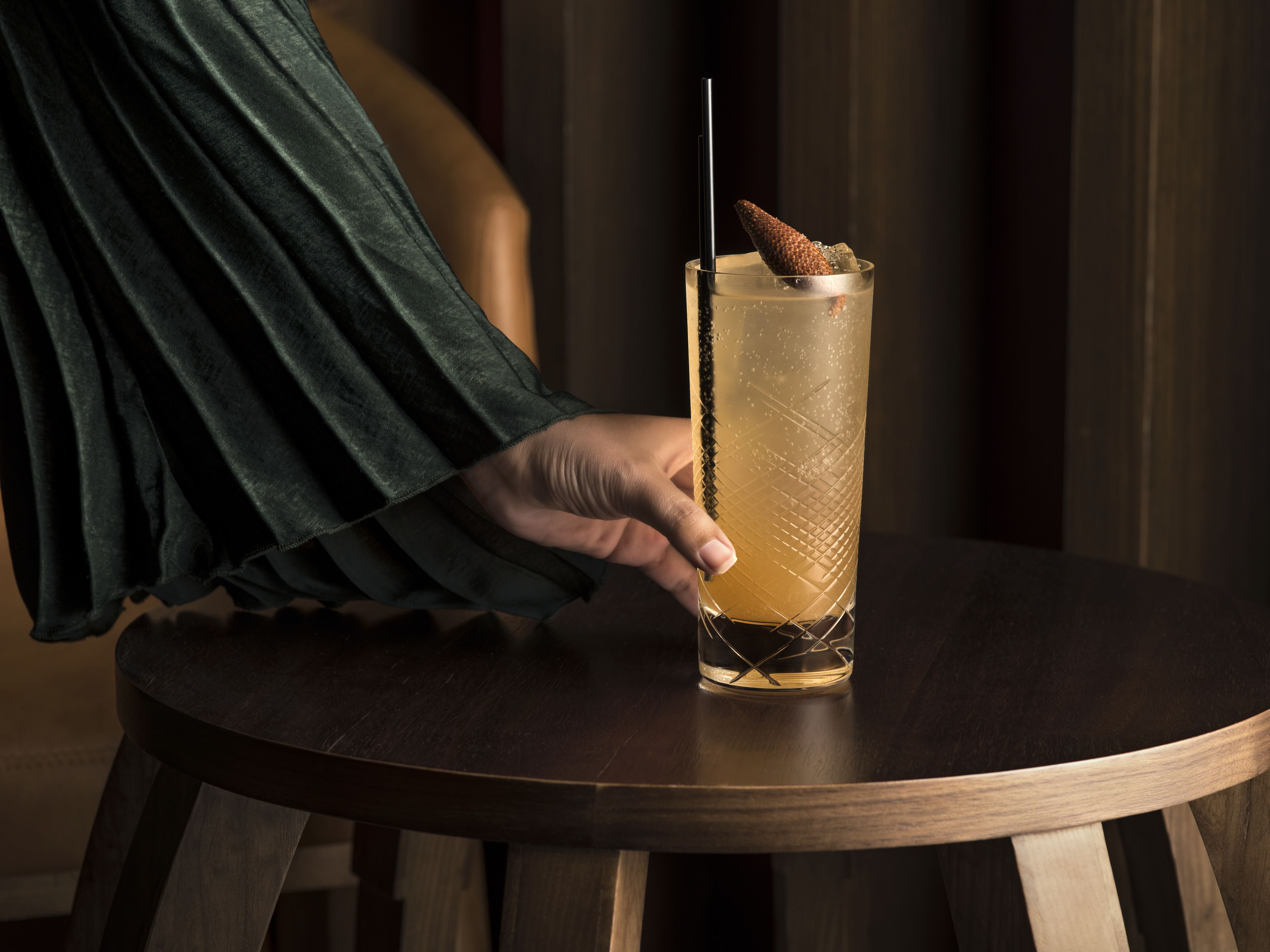 A person holding a tall glass with a straw and a decorative item, placed on a round wooden table.