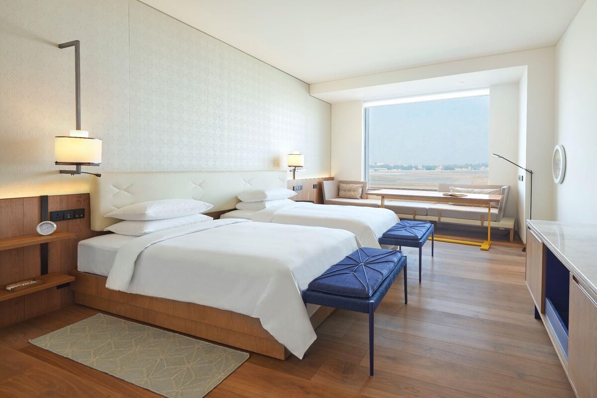 A hotel room with two beds, a desk, a chair, a TV, and a view of the airport.