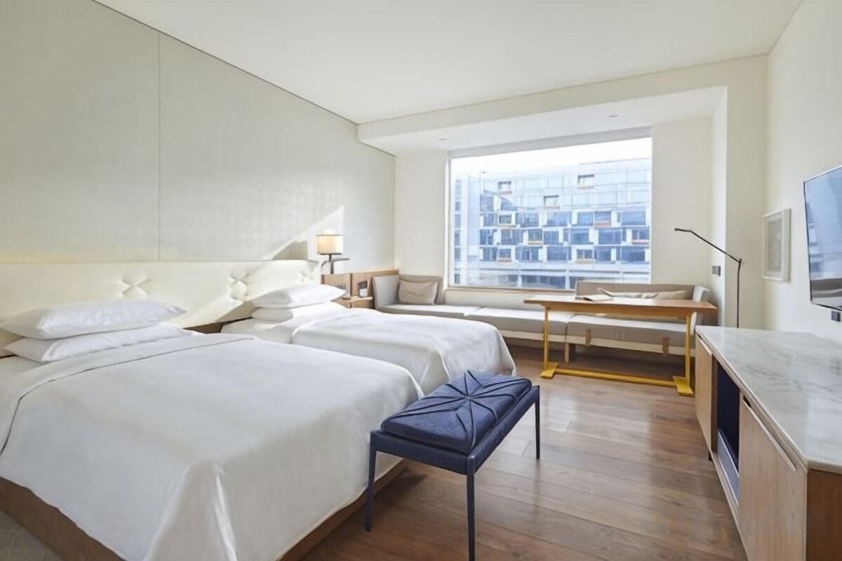A hotel room with two beds, a desk, a window with a city view, and a TV.