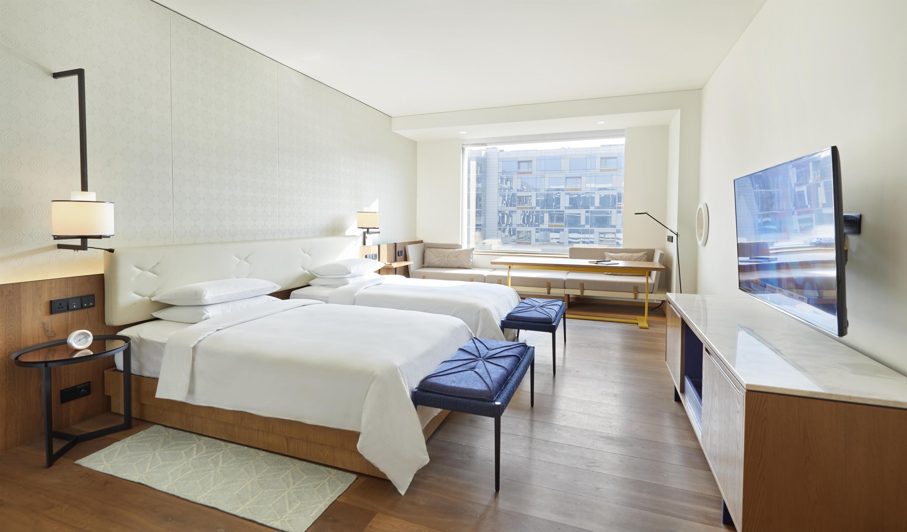 A hotel room with two beds, a sofa, a TV, and a window with a city view.