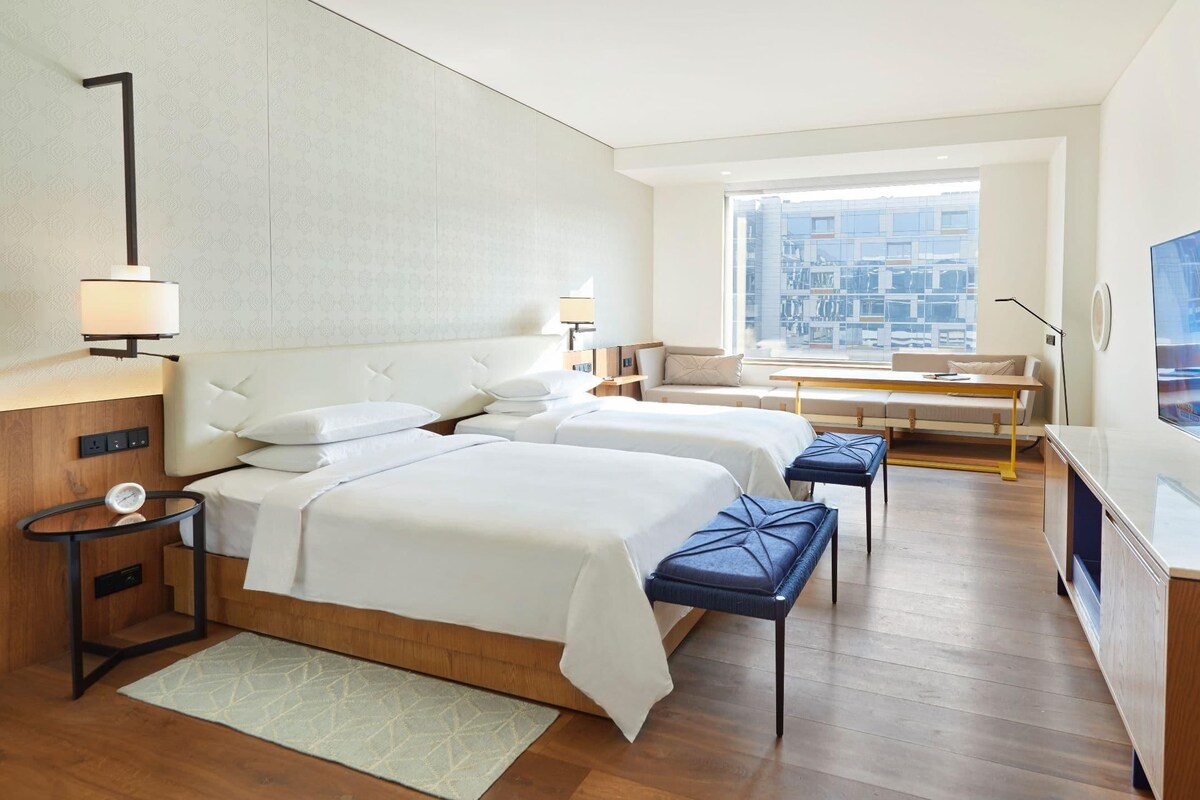 A hotel room with two beds, a desk, a TV, and a large window with a city view.